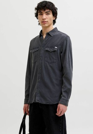 Jeune homme portant une chemise noire délavée à boutons avec des poches poitrine, tenant une poignée de sac noir sur un fond clair uni.