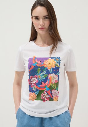 Jeune femme portant un t-shirt blanc avec un imprimé floral coloré et perroquet, associé à un pantalon bleu clair, debout devant un fond uni.