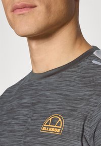 Grå kortermet skjorte med et subtilt teksturert mønster. Har en orange Ellesse-logo på venstre bryst og rund hals.