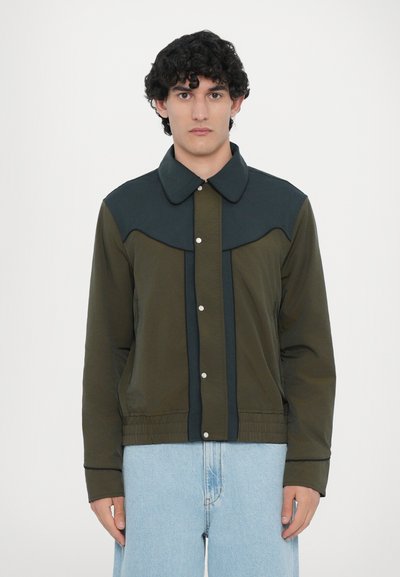 Veste vert olive avec un col vert foncé, panneaux contrastants, boutons-pression et poignets élastiques. Dispose d'une coupe droite et d'une texture lisse.