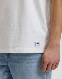 Λευκό βαμβακερό μπλουζάκι με ίσια γραμμή, διακοσμημένο με μια μικρή μπλε ετικέτα με το λογότυπο Lee στο κάτω στρίφωμα. Συνδυασμένο με ανοιχτό μπλε τζιν παντελόνι.