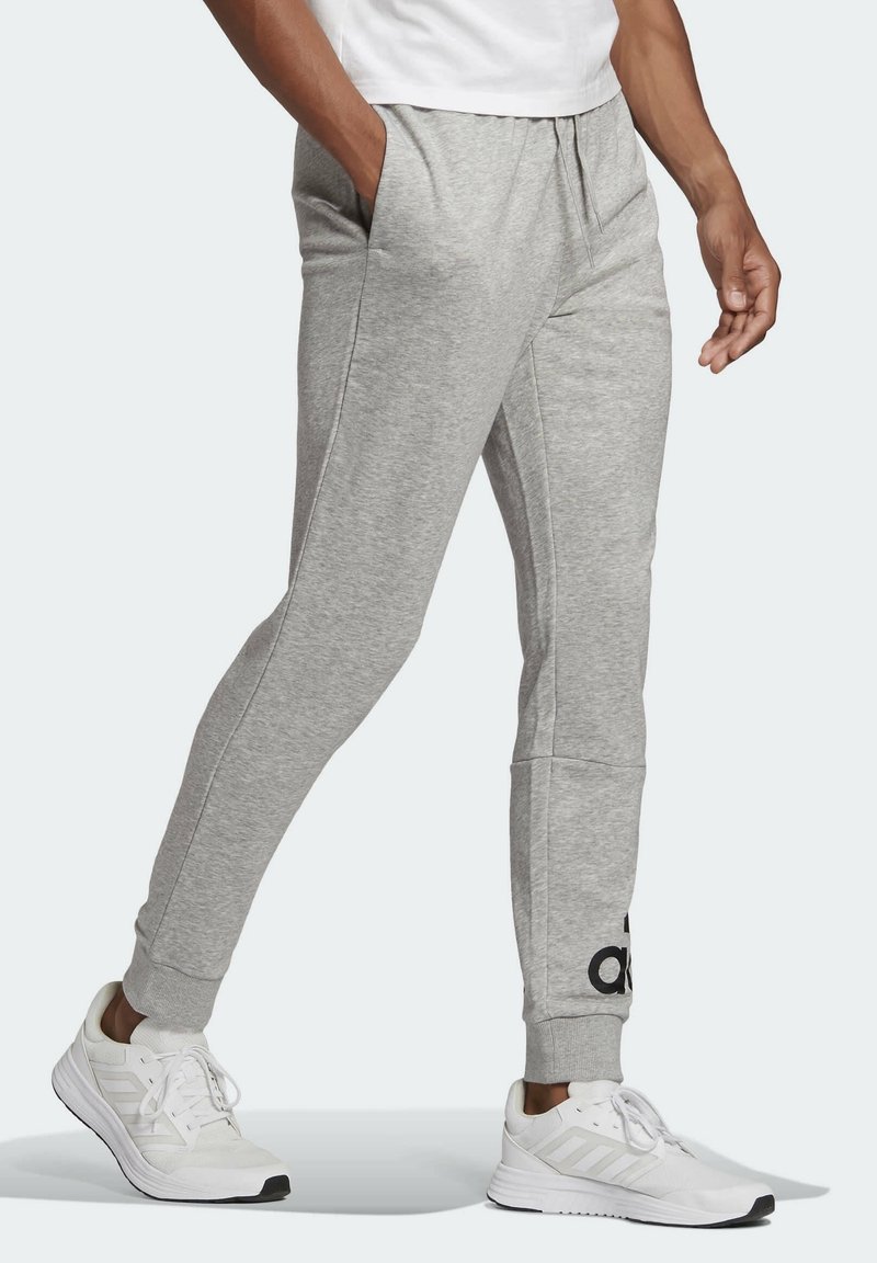 Grå joggingbukser lavet af blødt materiale, med sidelommer, tætsiddende ben og et sort logo nær kanten. Parret med hvide sneakers.