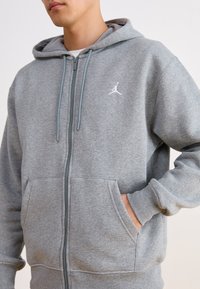 Felpa grigia con zip realizzata in morbido materiale felpato. Presenta una tasca a marsupio, cappuccio con cordino e logo bianco sulla parte sinistra del petto.