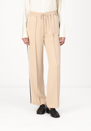 Beżowe spodnie o szerokich nogawkach z elastycznym pasem i czarno-białymi paskami po bokach, noszone z czarnymi mokasynami i kremową bluzką.