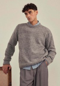 Pull en laine gris avec un col rond, présentant une finition douce et texturée. Porté sur une chemise à col rayée bleu clair, assorti à un pantalon gris.