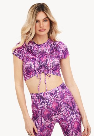 Mujer que lleva un top corto fruncido con estampado en tonos morado y rosa, combinado con leggings a juego de cintura alta, de pie con una mano a un lado.