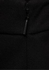 Mustan värinen kangas, jossa on vinoraitainen pinta ja vetoketju. Vetoketju on metallinen, matta viimeistelyllä, ja se sulautuu saumattomasti muotoiluun.