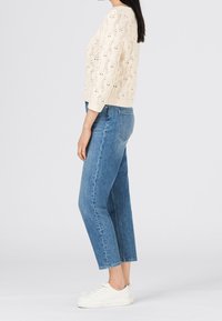 Pull en crème tricoté avec un motif en dentelle, associé à un jean taille haute bleu clair et des baskets blanches. Vue latérale montrant l'ajustement et le design.