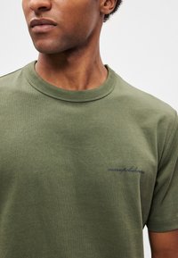 Oliivinvihreä puuvillainen t-paita pyöreällä kaula-aukolla, jossa on rintakehässä pieni sininen kirjailtu logo ja pehmeä, sileä pinta.