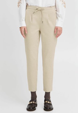 Beige tapered trousers with a drawstring waist, pleated front, and side pockets, paired with dark shoes and textured socks.
