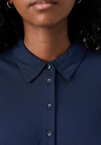 Tumši zili krekls ar apkakli, kas piedāvā pogu priekšpusi, ar sešām apaļām pogām un gludu, nedaudz teksturētu audumu.