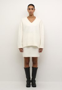 Witte gebreide v-halstrui met een ruitpatroon, gecombineerd met een bijpassende rok. Zwarte hoge laarzen met gespen maken de outfit af.