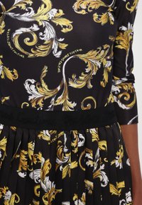 Black dress with bold yellow and white floral patterns. Features a pleated skirt and a textured black waistband with embossed brand name.