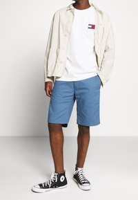 Bílé tričko s barevným logem, béžová košová bunda, modré kraťasy a černé vysoké tenisky. Textury látek se liší napříč jednotlivými kousky.