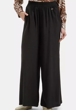 Kobieta nosząca luźne czarne spodnie z szerokimi nogawkami i elastycznym pasem, ręce w kieszeniach, w połączeniu z wzorzystym długim rękawem i beżowymi butami.