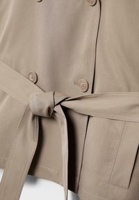 Tessuto di cappotto trench beige con cintura annodata, quattro bottoni e cuciture della tasca visibili.