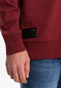 Bordeauxfarbenes Sweatshirt mit gerippten Bündchen, ausgestattet mit einem schwarzen Lederpatch am Saum. Weiche Textur, taillierte Passform, kombiniert mit blauen Jeans.