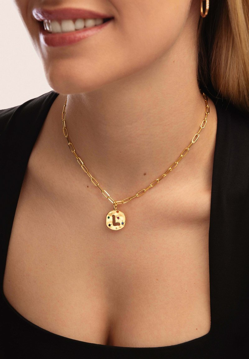 Collier en or avec des maillons allongés et un pendentif circulaire mettant en valeur une lettre et des accents colorés. Surface texturée avec une finition polie.