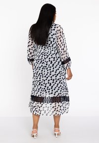Langes, transparentes Kleid mit einem weißen Hintergrund, schwarzen und braunen abstrakten Flecken, mit einer Spitzenkante und langen Ärmeln.