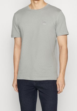 Grå bomuld t-shirt med korte ærmer, rund halsudskæring og et lille mærke på venstre bryst. Båret sammen med mørkeblå jeans.