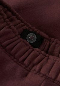 Suurendatud vaade maroonist elastsele vöötmele, millel on reguleeritav must rihm ja ümar selge nupp kangal.