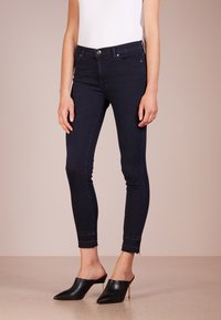 Dunne donkerblauwe jeans met een hoge taille, voorzien van riemlussen en subtiele vervaging. De zoom is lichtjes rafelig. Gepaard met zwarte muilen.