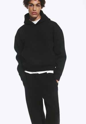Jongere met krullend haar die een zwarte hoodie en bijpassende zwarte broek draagt, staat met de handen in de zakken tegen een effen achtergrond.