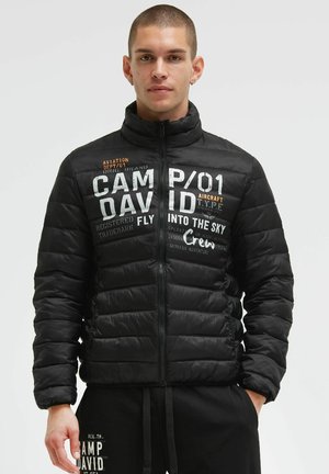 Young man with short hair wearing a black padded jacket and matching black pants with white text design.