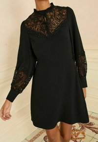Črna obleka z dolgimi rokavi in čipkastim detajlom na ovratniku in rokavih. Mehka tkanina, oprijet pas in razširjen krilo.