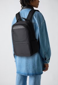 Sac à dos noir avec une texture lisse, en forme rectangulaire et avec des bretelles ajustables. Comprend une poche zippée à l'avant et un logo de marque subtil sur le côté.