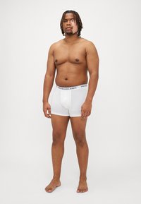 Fehér pamut boxer alsó fekete márkázott derékpánttal, testhezálló kivitelben, combközép hosszal, mezítláb álló személy viseli.