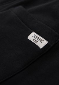 Zwart stof met een gladde textuur heeft een genaaide witte label met de tekst: "JOUW ENIGE LIMIET IS JE GEEST," merknaam: "OMBRE," en "SINCE 1985."