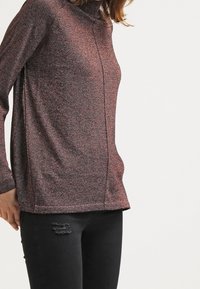 Woman wearing a brown textured long-sleeve turtleneck sweater and black ripped skinny jeans, standing against a white background.