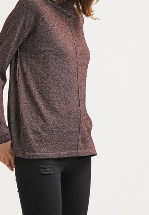 Eine Frau trägt einen braunen strukturierten Langarmrollkragenpullover und schwarze, zerrissene Skinny Jeans und steht vor einem weißen Hintergrund.