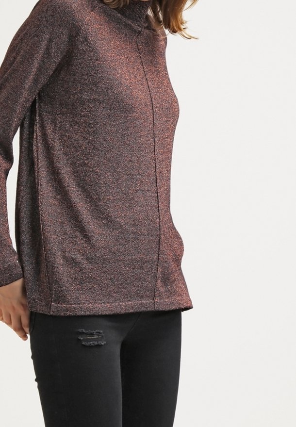 Woman wearing a brown textured long-sleeve turtleneck sweater and black ripped skinny jeans, standing against a white background.