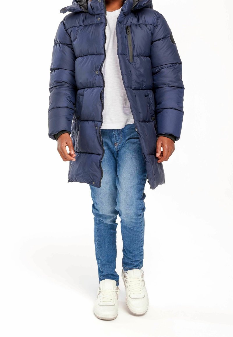 Personne portant une doudoune bleu marine, un t-shirt blanc, un jean bleu et des baskets blanches, debout sur un fond blanc.
