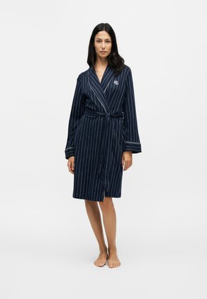 Femme debout pieds nus portant une robe de chambre bleu marine à fines rayures avec ceinture à la taille et logo "RL" sur la poitrine sur fond uni.