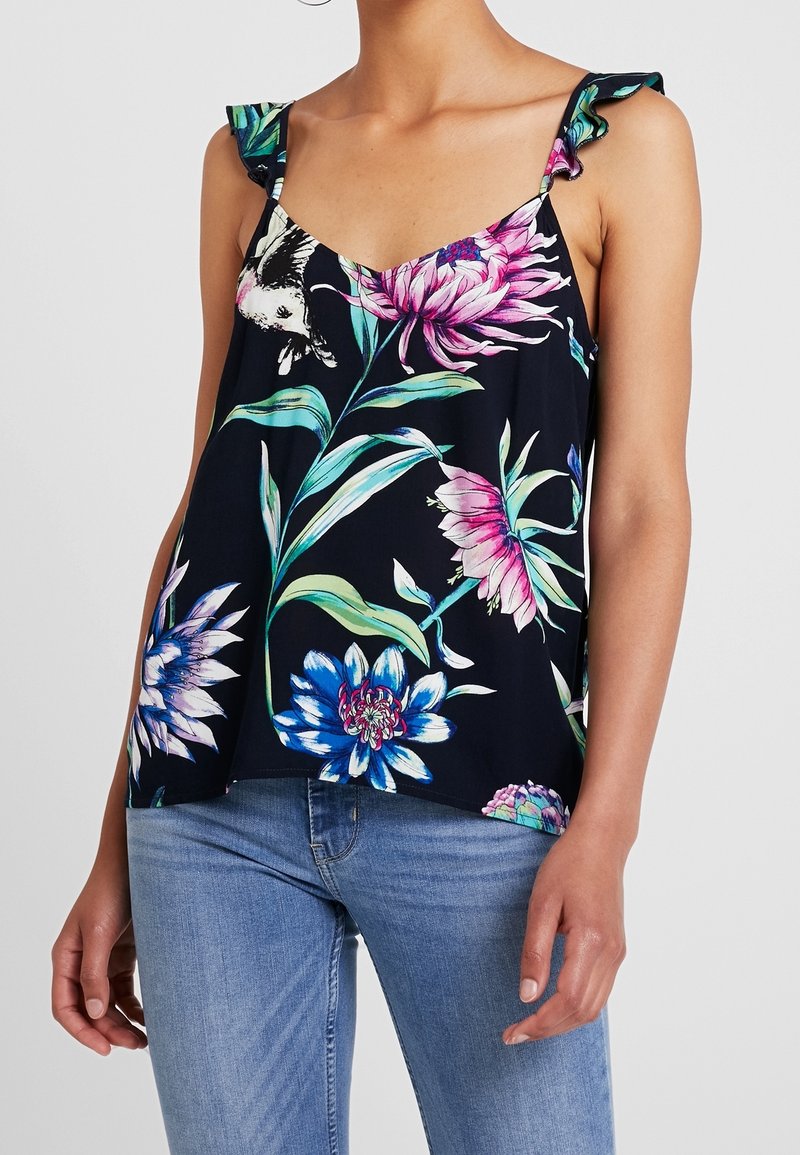 Femme portant un haut sans manches à motifs floraux foncés avec des bretelles à volants et un jean bleu, montrant le ventre et le haut des cuisses.