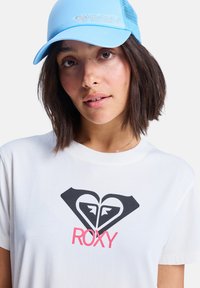 En ung kvinne med skulderlangt hår har på seg en hvit t-skjorte med en svart og rosa Roxy-logo, supplert med en blå caps.