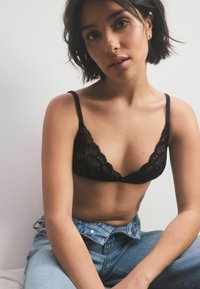 Zwarte kanten bralette met een driehoekige vorm, versierd met delicate florale patronen en verstelbare bandjes, gecombineerd met hooggeknoopt denim jeans.