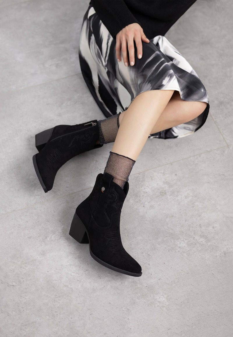 Botines de ante negros con punta afilada y tacón ancho. Presentan costuras decorativas y cierre lateral con cremallera. Calcetines de tobillo grises texturizados visibles.