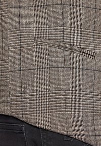 Brązowa tkanina w kratkę o teksturowej powierzchni, z małą kieszonką i widocznym szyciem, w połączeniu z czarną tkaniną poniżej.