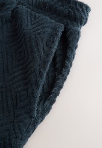 Mörkblå handduk med texturerat geometriskt mönster, mjuk, plyschig yta och vågig kantdetalj, vikt på en ljus bakgrund.