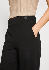 Pantalon noir en tissu texturé, avec une poche latérale et un bouton noir à la taille, présenté partiellement avec un haut au-dessus.
