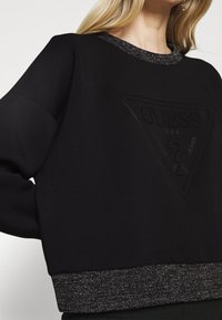 Czarna bluza z fakturowanym logo w kształcie trójkąta, błyszczącym dekoltem i lśniącym wykończeniem. Miękki materiał o luźnym kroju.