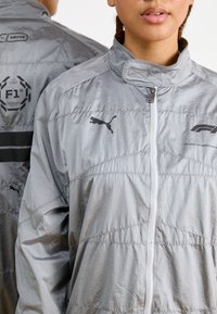Sølv polstret jakke med høy krage, glidelås foran og svarte logo detaljer. Teksturt stoff med vertikale sømmer og synlig sport branding.