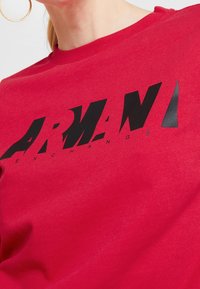T-shirt di cotone rosso con un grande logo nero caratterizzato da un font moderno e un design angolare. Collo a giro e texture liscia.