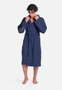 Accappatoio blu navy realizzato in tessuto strutturato, con cappuccio, design avvolgente, vita con cintura e due tasche frontali con dettaglio del logo sulla manica.