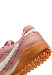Sneaker Nike en rose clair avec des accents blancs, matériau lisse, bout arrondi, et semelle en caoutchouc orange présentant des motifs de traction.