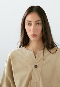 Beige Wildlederoberteil mit rundem Ausschnitt, das über einen einzelnen Knopfverschluss am Kragen und weite Ärmel verfügt. Weiche Textur, minimales Design.